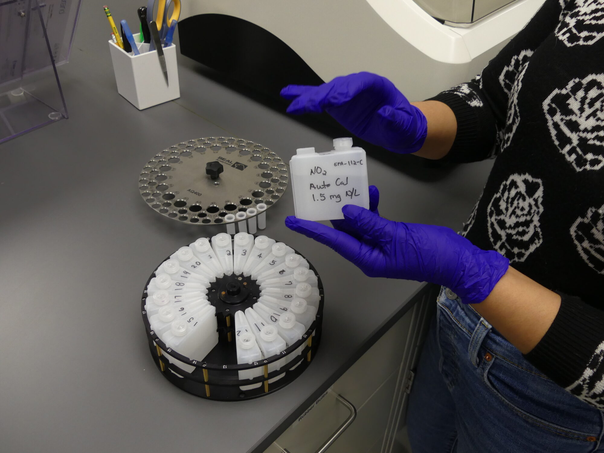 Wearing a pair of purple gloves, Elena Colon places reagents and standards into the analyzer at The John and Daria Barry Foundation Laboratory in Larchmont. 