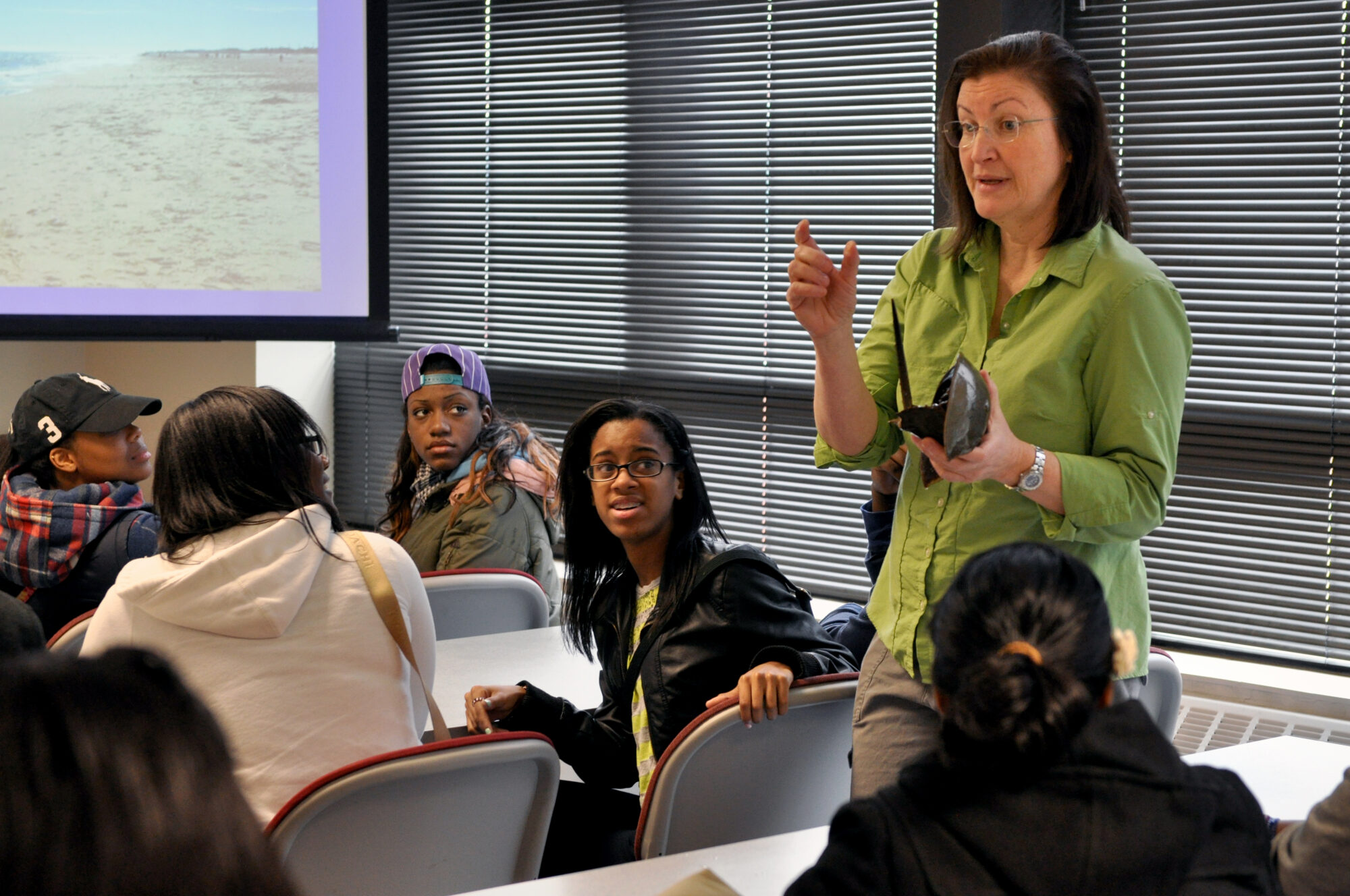 Several high school students sit at tables and turn to face and listen to a woman wearing a green shirt addressing the group. 