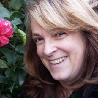 A middle-aged white women poses against a fence and smiles in a selfie taken next to a large pink flower.