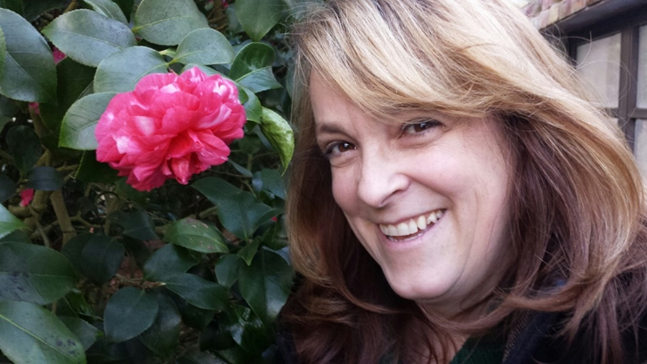 A middle-aged white women poses against a fence and smiles in a selfie taken next to a large pink flower.