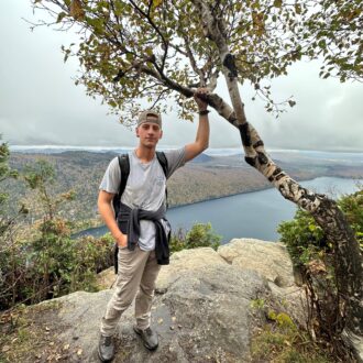 A white man poses next to a tree on top of a mountain.