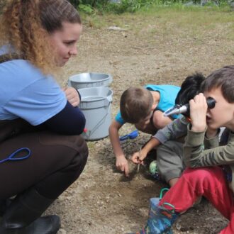 A young woman wearing waders crouches down to speak with a group of three young boys looking at research tools.