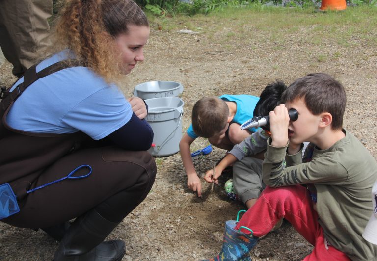 A girl wearing waders crouches down to a group of young boys looking at salinity refractometers.