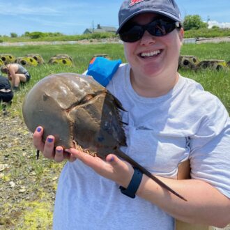 A woman in sunglasses and a white t-shirt holds up a horseshoe crab and smiles.