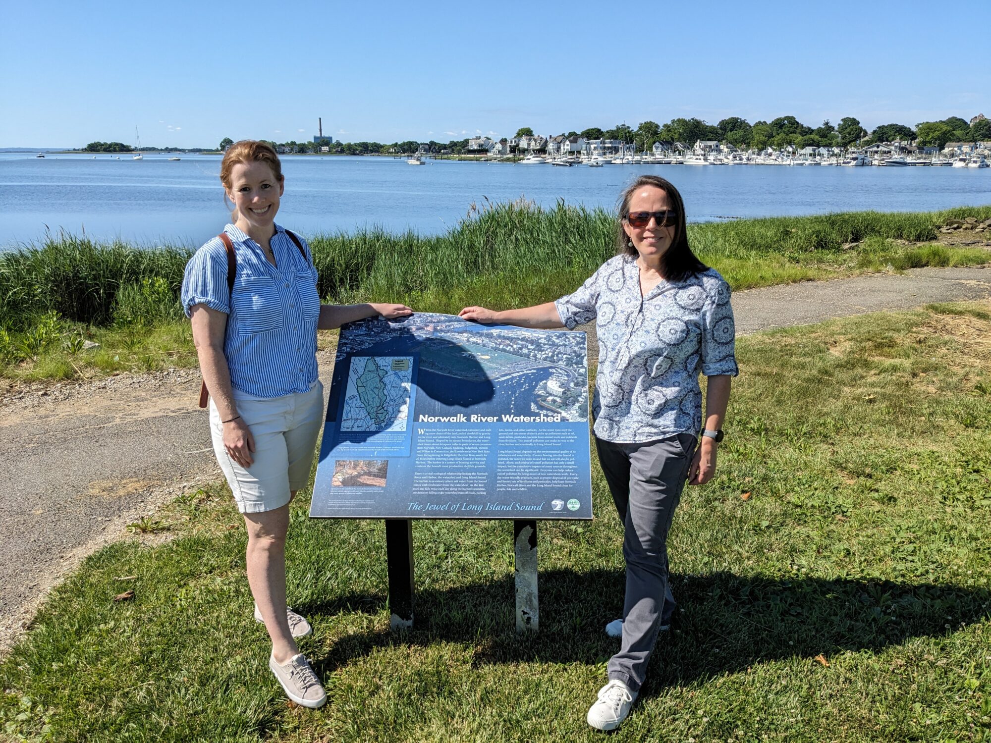 Two women stand next to a large blue sign in a waterfront park. 