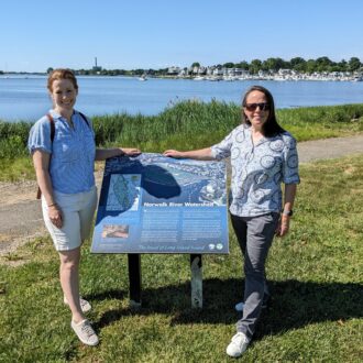 Two women stand next to a large blue sign in a waterfront park.