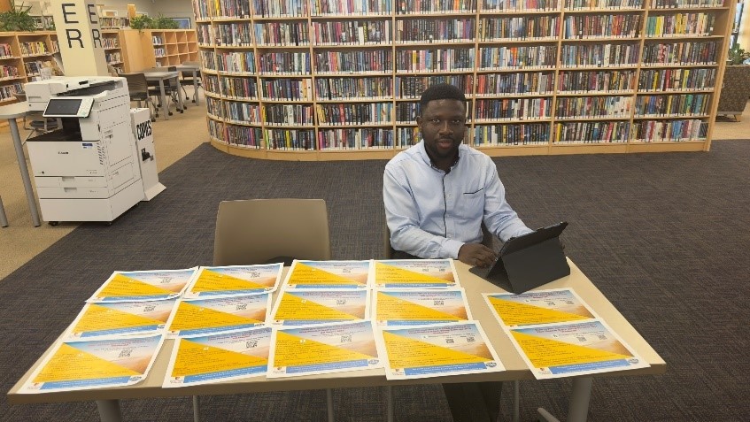 A black man sits at a table in a library with yellow fliers.