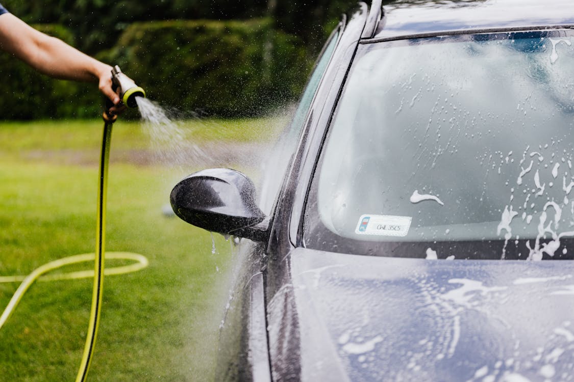 White man washes a car on grass with a garden hose. 