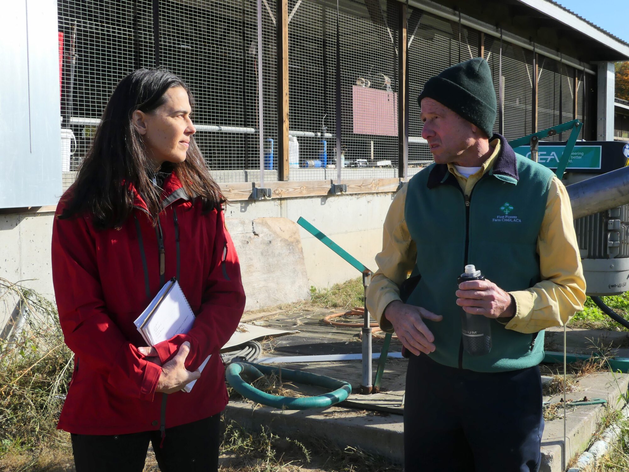 A young woman with brown hair and a red jacket looks over at a white man with a green hat and green vest. They stand together in front of a barn and talk. 