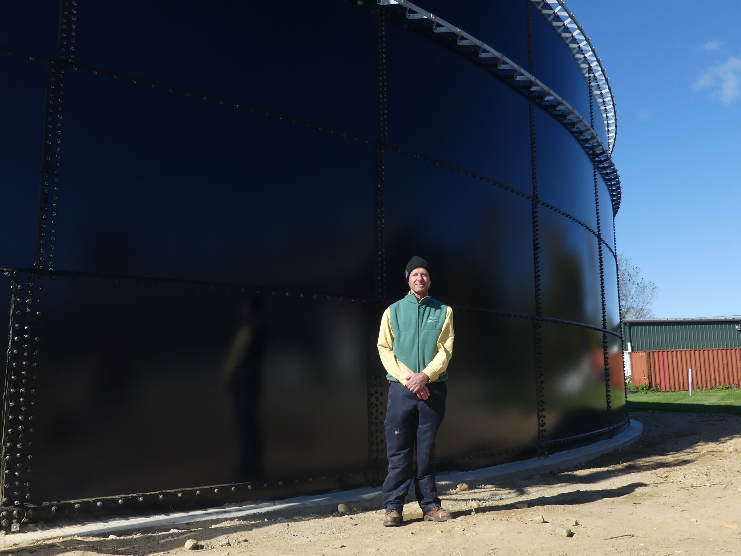 A white man stands in front of a large black manure storage tank on a farm. He is smiling and has a hat and vest on. 