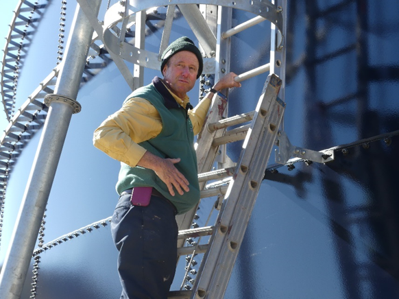 An older white man on a ladder leaned against a large black structure. He wears a hat and a vest. 