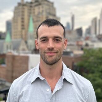 Professional headshot of a brunette white man in a blue shirt. A city skyline in the background.