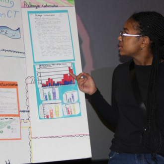 A young girl with braids presents a science project on pathogen contamination, featuring charts and handwritten notes on a display board.