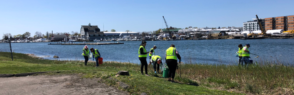 People in safety vests and cleaning tools picking up trash along a shoreline.